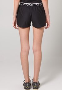 Czarne sportowe spodenki o dopasowanym kroju, z białym pasem w talii z napisem. Noszone z czarnymi i żółtymi butami do biegania.