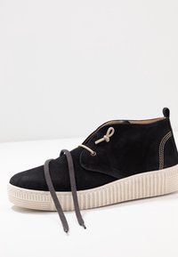 Černá semišová bota s kulatou špičkou, béžovou rýhovanou podrážkou, šedými tkaničkami a hnědými akcenty, s háčkem pro bezpečné zapínání.