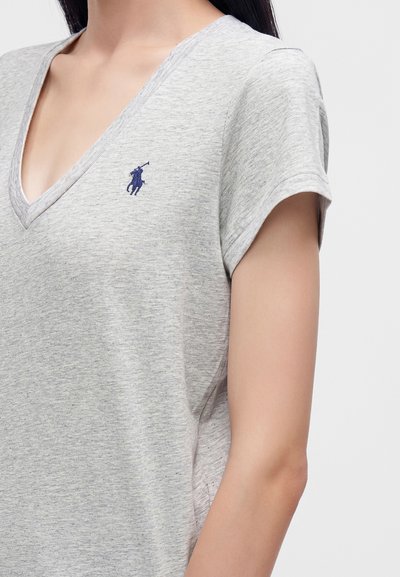 Γυναίκα που φοράει ανοιχτό γκρι T-shirt με V-λαιμόκοψη και μικρό σκούρο μπλε λογότυπο παίκτη πόλο στο αριστερό στήθος, κοντά μανίκια.