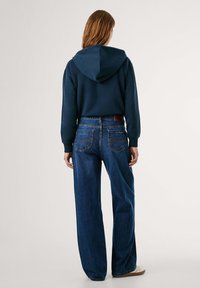 Persoon in een marineblauwe hoodie en hooggewaist wijde blauwe jeans, staand met de rug naar de camera tegen een effen lichte achtergrond.
