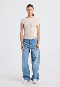 Nuori nainen seisoo päällä valkoinen pitsinen lyhythihainen paita, väljät vaaleansiniset farkut ja valkoiset lenkkarit yksinkertaista taustaa vasten.