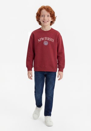 Smilende rødhåret dreng går fremad iført en rød New Jersey sweatshirt, blå jeans og hvide sneakers mod en hvid baggrund.