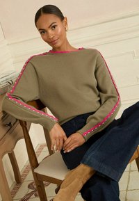 Olíva zöld pulóver rózsaszín díszítéssel az ujjak mentén, kötött textúrával és kerek nyakkivágással. Sötétkék farmernadrággal és bézs csizmával párosítva.