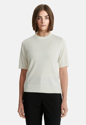 Mujer con cabello castaño liso, vestida con un suéter blanco de manga corta y pantalones negros, de pie frente a un fondo claro y sencillo.