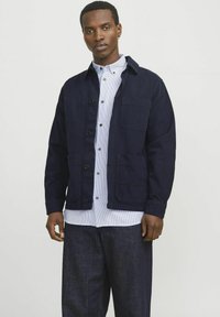 Navy blue jacket with a button-up front, two chest pockets, and a collared neck. Worn over a light blue striped shirt. Dark denim trousers.