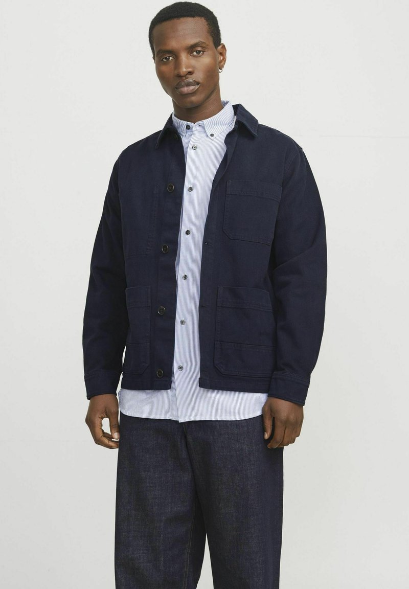 Navy blue jacket with a button-up front, two chest pockets, and a collared neck. Worn over a light blue striped shirt. Dark denim trousers.