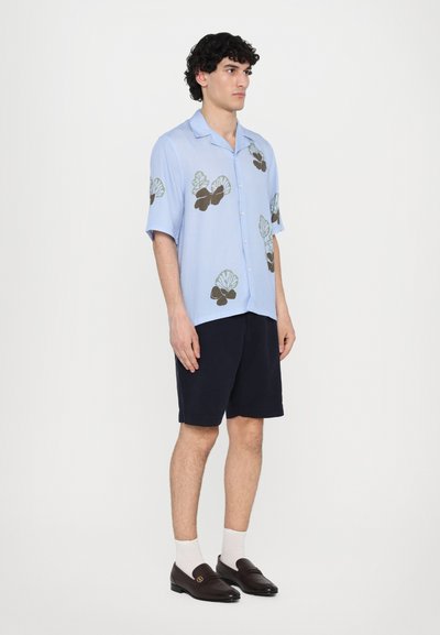 Lyseblå kortermet skjorte med mørkegrønne og brune skjelltrykk, kombinert med marineblå shorts og brune loafers. Hvite sokker er synlige.