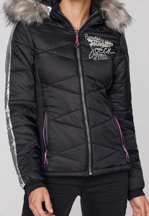 Femme portant une veste matelassée noire avec des bandes argentées sur les manches, une capuche bordée de fourrure, des fermetures éclair roses et un texte brodé sur la poitrine.
