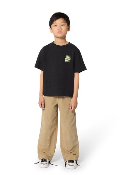 Αγόρι στέκεται φορώντας μαύρο γραφικό T-shirt, μπεζ φαρδιά παντελόνια και λευκά αθλητικά παπούτσια με μαύρα κορδόνια σε λευκό φόντο.