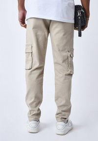 Pantaloni cargo beige con tasche laterali, vestibilità affusolata, realizzati in tessuto leggero. Abbinati a sneaker bianche, con una maglietta bianca semplice visibile sopra.