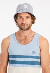 Blauer Bucket Hat aus leichtem Stoff, mit einem umklappbaren Schirm und einem kleinen Logo-Patch. Getragen mit einem hellblauen Tanktop.