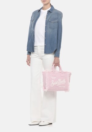 Persona che indossa una camicia di denim blu, una t-shirt bianca e pantaloni, che tiene in mano destra una borsa a tracolla rosa con scritta bianca "Saint Barth".