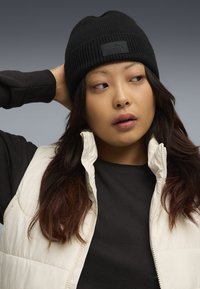 Black ribbed knit beanie with a textured finish and a branded tag, worn with a white puffer vest over a black shirt.