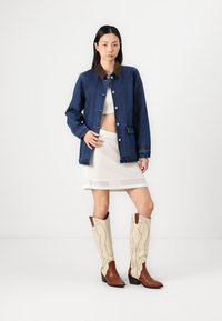 Jeansjakke med brun krage, hvit cropped topp, teksturert hvit skjørt, og brune og kremfargede cowboystøvler med dekorativt design.