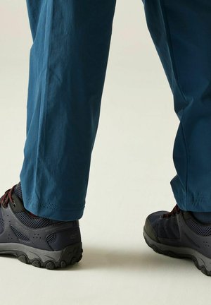 Persona con zapatos de senderismo azul marino y pantalones azules para exteriores de pie sobre una superficie clara.