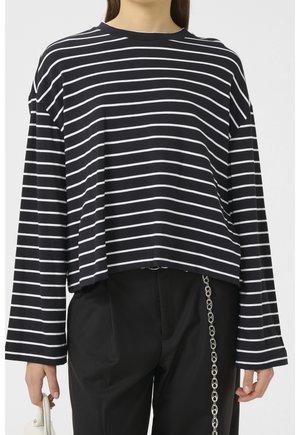 Persoon draagt een zwart-wit horizontaal gestreept shirt met lange mouwen en een zwarte broek met een zilveren kettingaccessoire.