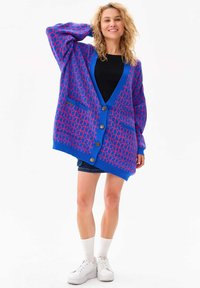 Blauwe cardigan met een roze geometrisch patroon, knoopsluitingen, twee voorkruiszakken, lange mouwen en een relaxte pasvorm. Gstyled over een zwart top.
