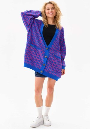 Blauwe cardigan met een roze geometrisch patroon, knoopsluitingen, twee voorkruiszakken, lange mouwen en een relaxte pasvorm. Gstyled over een zwart top.