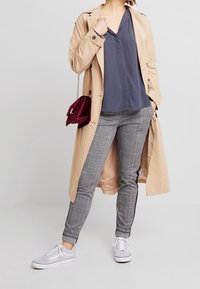 Beige trenchcoat över en marinblå blus, grå rutiga byxor och grå sneakers. Accessoarer inkluderar en vinröd handväska med kedjesnodd.