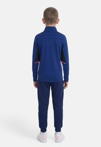 Παιδί που στέκεται με την πλάτη στραμμένη, φοράει μπλε φόρμα με λευκές και ροζ λεπτομέρειες και λευκά αθλητικά παπούτσια Nike με ροζ λογότυπα.