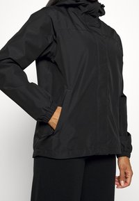 Chaqueta negra impermeable con capucha, que presenta puños elásticos, bolsillos frontales y un diseño aerodinámico. Textura suave y mate, sin patrones visibles.