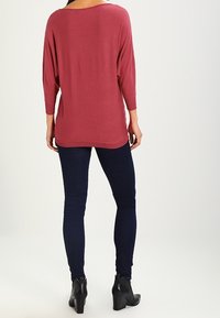 Maglietta a maniche lunghe di colore bordeaux con una vestibilità ampia e scollatura larga. Abbinata a jeans skinny scuri e stivaletti, il tutto su uno sfondo semplice.