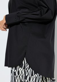 Schwarzes Langarmshirt mit abgerundetem Saum, ausgestattet mit geknöpften Manschetten und einer glatten, gewebten Stoffstruktur. Darunter ist ein gemusteter Rock sichtbar.