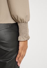 Manches longues beiges avec poignets froncés et détail subtil de volants, associées à un pantalon noir texturé, main détendue sur le côté.