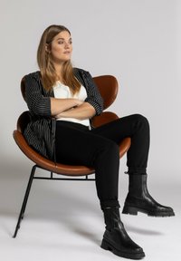 Černé kotníkové boty s masivními podrážkami; modelka v pruhované černobílé bundě a černých kalhotách, sedící v hnědé, mušle tvarované židli.