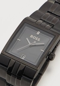 Μαύρο ορθογώνιο ρολόι χειρός BOSS με λουράκι από μεταλλικές συνδέσεις, ασημένιους δείκτες ώρας και λεπτών, ένδειξη ημερομηνίας που δείχνει 23, και ελάχιστους δείκτες ώρας.