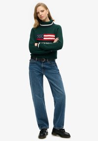 Pull en tricot vert avec motif du drapeau américain, associé à un jean bleu délavé à jambes larges. Une ceinture noire et des chaussures noires complètent l'ensemble.