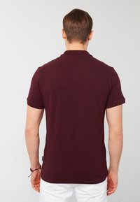 Burgundivärinen polo-paita näkyy takaapäin, siinä on lyhyet hihat, kaulus ja sileä pinta. Valkoinen etiketti näkyy sivussa.
