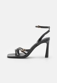 Black high-heeled sandals made of glossy leather. Features crisscross straps with gold studs and a buckle closure on the ankle strap.