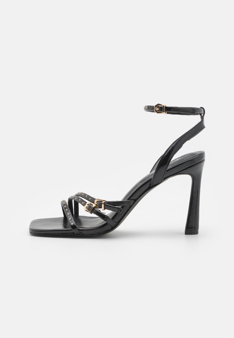 Black high-heeled sandals made of glossy leather. Features crisscross straps with gold studs and a buckle closure on the ankle strap.