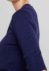 Tmavě modrý svetr s kulatým výstřihem a tříčtvrtečními rukávy. Rukávy jsou na lemu zdobeny texturovaným krajkovým vzorem. Měkký pletený materiál.
