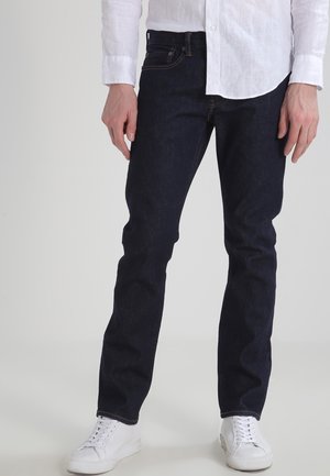 Mann trägt dunkelblaue Jeans, weißes Hemd mit Knöpfen und weiße Turnschuhe, steht vor einem schlichten, hellen Hintergrund.