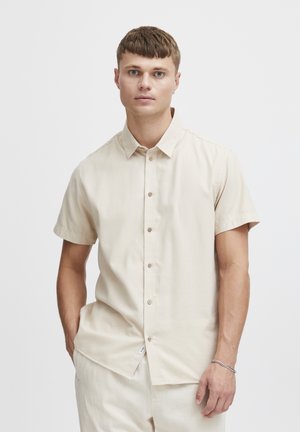 Camisa de botones de manga corta en color beige, elaborada con un tejido suave, que cuenta con un cuello, botones en la parte delantera y una etiqueta lateral sutil.