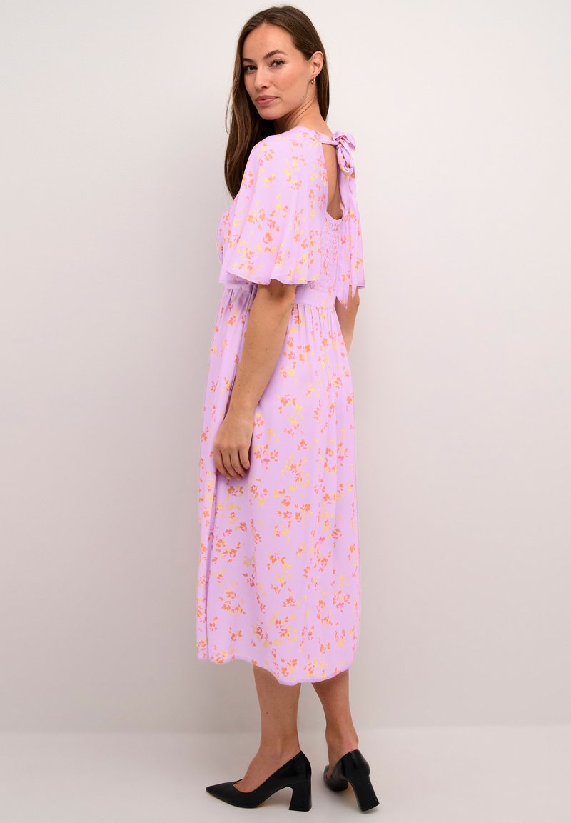 Robe longue à fleurs roses avec des manches courtes ondulées, un détail de nœud au dos et une coupe décontractée. Portée avec des talons noirs pointus.