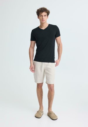 Mees seisab, kandes musta lühikeste varrukatega V-kaelusega särk, beeže lühikesed püksid ja beeže libisemisjalanõud ühtlasel heledal taustal.