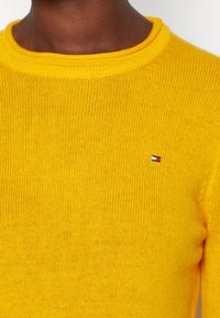 Pull jaune en tricot avec un col rond et des bordures côtelées. Présente un petit écusson logo en rouge, blanc et bleu sur la poitrine.
