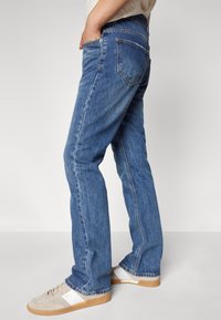 Blå denim jeans med rett passform, med baklommer og en lett falming. Brukt med beige og hvite sneakers på en grå bakgrunn.