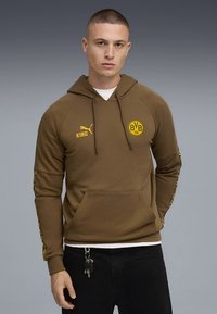 Hnedá mikina s kapucňou so šnúrkami, klokanie vrecko, žlté detaily loga a vzorované akcenty na rukávoch. Vyrobená z mäkkej tkaniny.