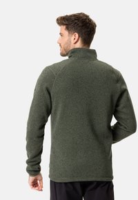 Jachetă din fleece verde cu guler înalt, material texturat și mâneci lungi, având cusături plate și un etichetă cu logo pe tivul de jos.