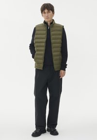 Mladenič nosi olivno zeleno puhovko čez črn pulover na zadrgo, črne hlače in črne škornje, ki stoji ob beli steni.