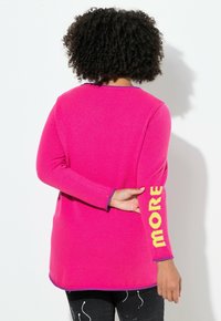 Person mit lockigem Haar, der einen leuchtend pinken Pullover mit gelbem "MORE"-Text auf dem Ärmel trägt und vor einer weißen Wand steht.
