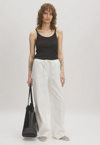 Zwarte geribbelde tanktop, witte linnen broek met koord, zwarte tote bag en geweven instap schoenen. Neutrale achtergrond benadrukt de kledingdetails.