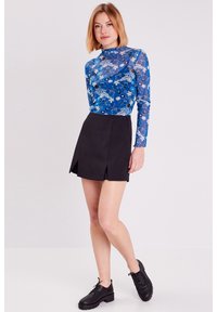 Haut bleu à manches longues avec un motif floral et une texture transparente, associé à une mini-jupe plissée noire. Le modèle porte des chaussures à lacets noires.