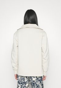 Světlý béžový svetr s rozšířeným límcem, spadlými rameny a žebrovanými manžetami. Texturovaná tkanina, jednoduchý design a volný střih.