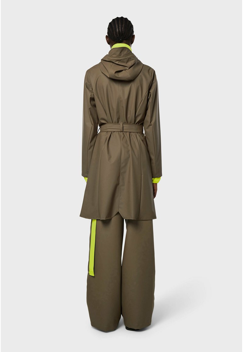 Persona con capelli intrecciati che indossa un cappotto con cappuccio marrone con cintura e pantaloni larghi con striscia gialla neon sul lato, vista di spalle.