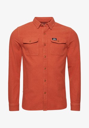 Roestkleurig oranje overhemd met lange mouwen, knopen, twee borstzakken met kleppen en een klein zwart merkje op de rechterzak.